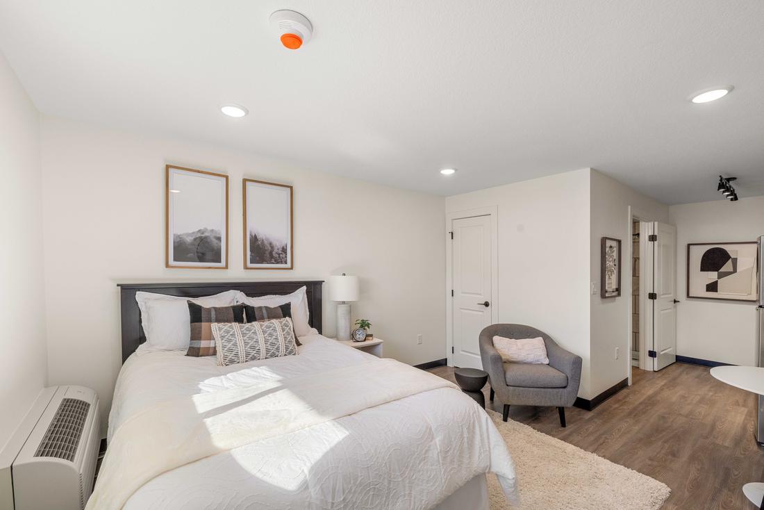 A staged studio apartment with a queen bed, framed prints, a lounge chair and wide-plank flooring.
